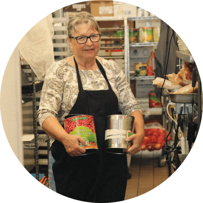 A woman wearing glasses and an apron is standing in a kitchen, holding two cans of food. She has a patterned shirt and appears to be in a food preparation area with shelves of ingredients visible in the background.