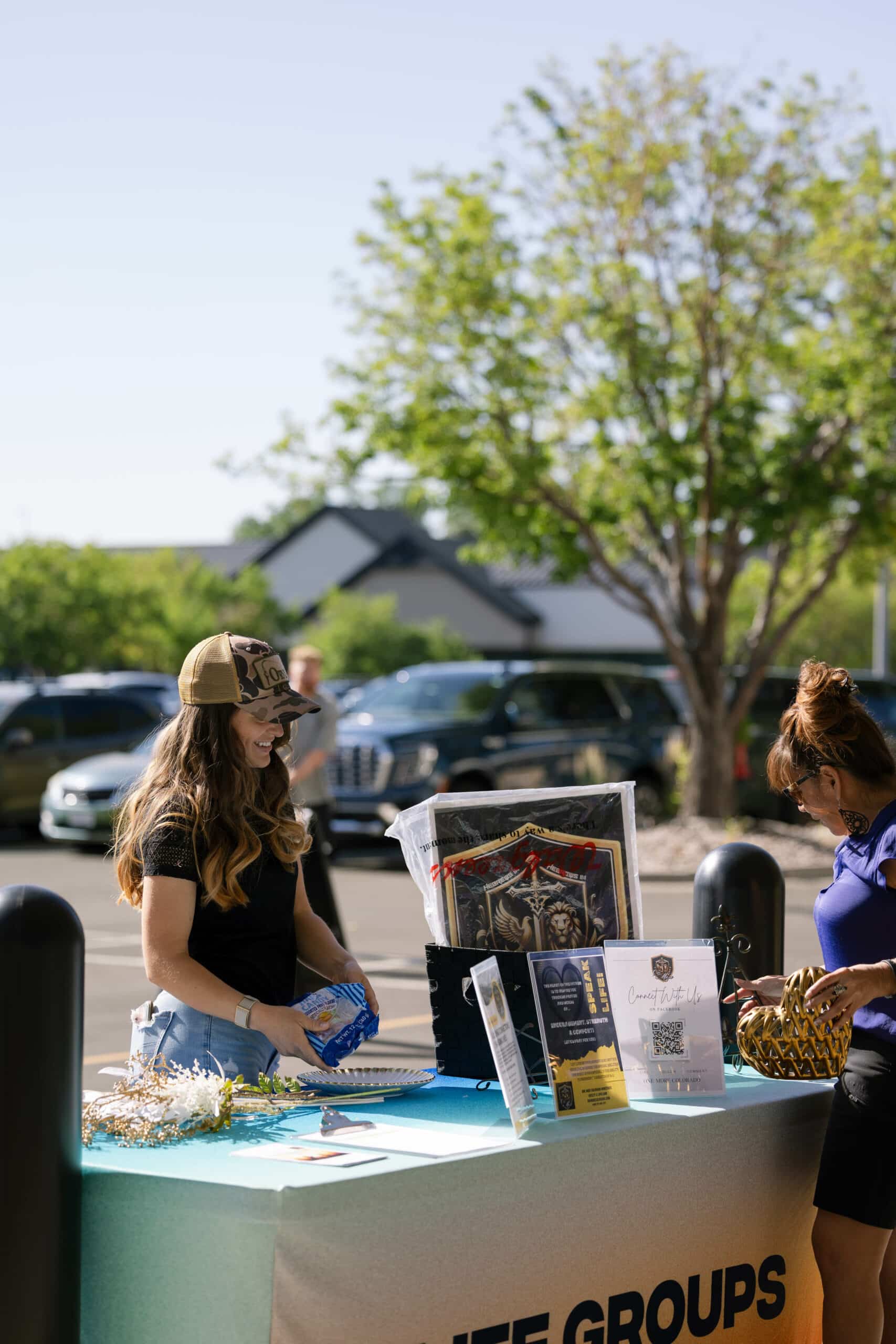 A woman wearing a cap and a black top stands behind a table with informational materials and a display. Another woman is beside her, reaching for a decorated item. In the background, parked cars and trees are visible.