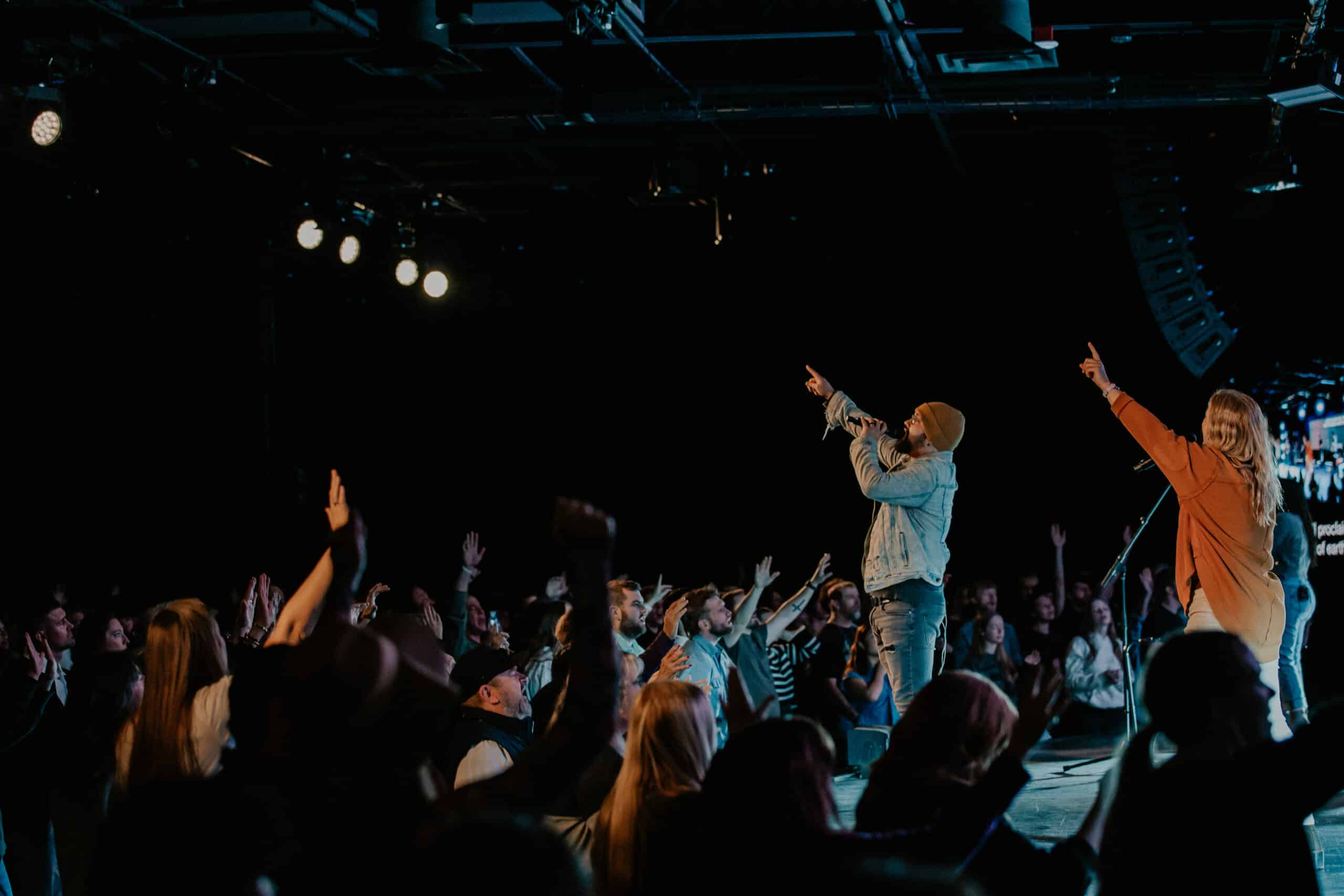 A singer in a denim jacket performs on stage with a microphone, while a second performer stands nearby. The audience is seen cheering and raising their hands in response, with stage lights illuminating the scene.