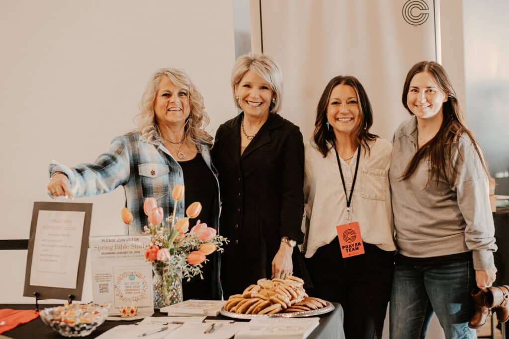Four women stand together at a table with baked goods. One woman wears a blue plaid shirt, another is dressed in black, while the others wear gray and white tops. The table features a floral arrangement and a platter of cookies, with informational materials visible.