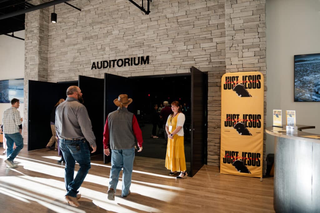 Three individuals are shown in a well-lit lobby area with a large sign reading "AUDITORIUM" above double doors leading into the auditorium. To the right, a banner displays the text "Just Jesus" next to a reception desk.