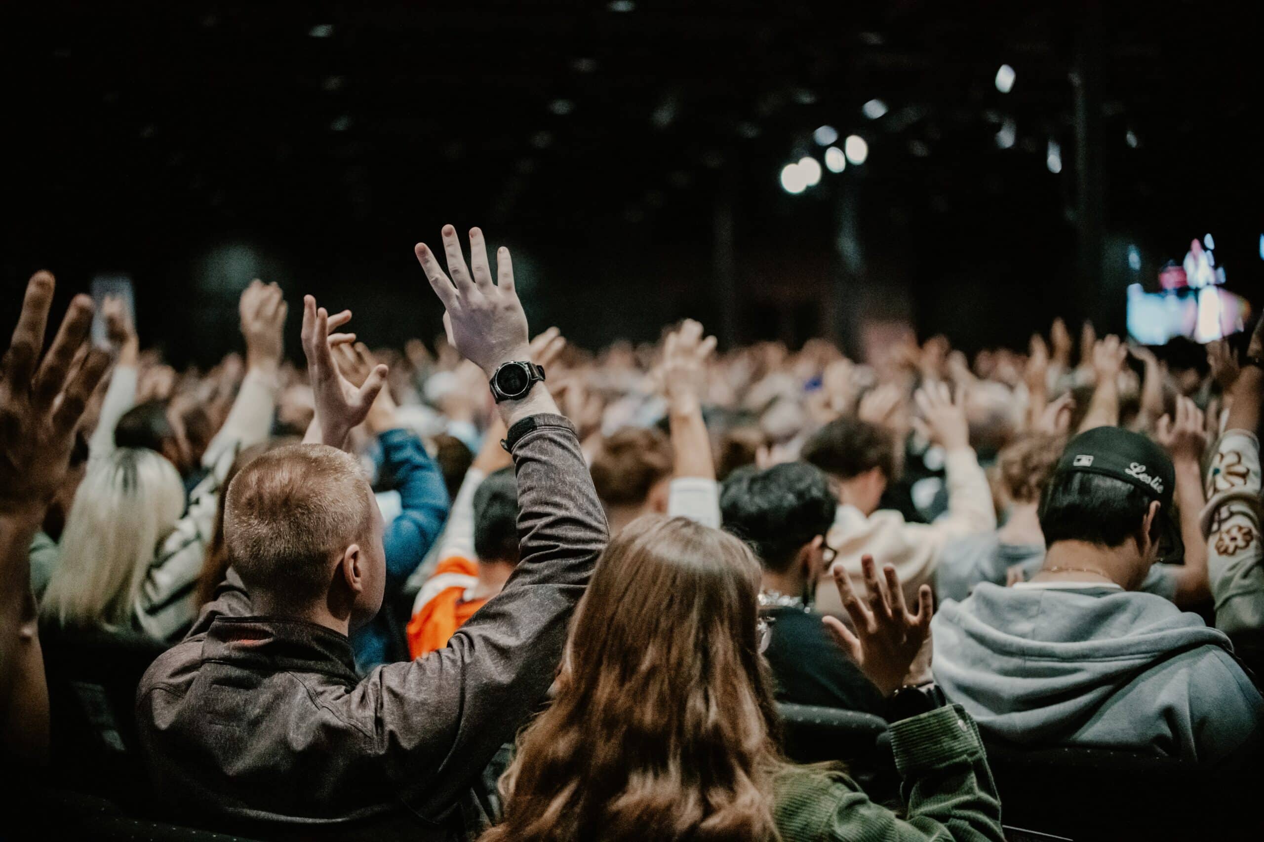 A large audience with many individuals raising their hands in a dimly lit auditorium. The crowd's focus appears directed towards a stage or presenter, with a variety of styles in clothing among the attendees.