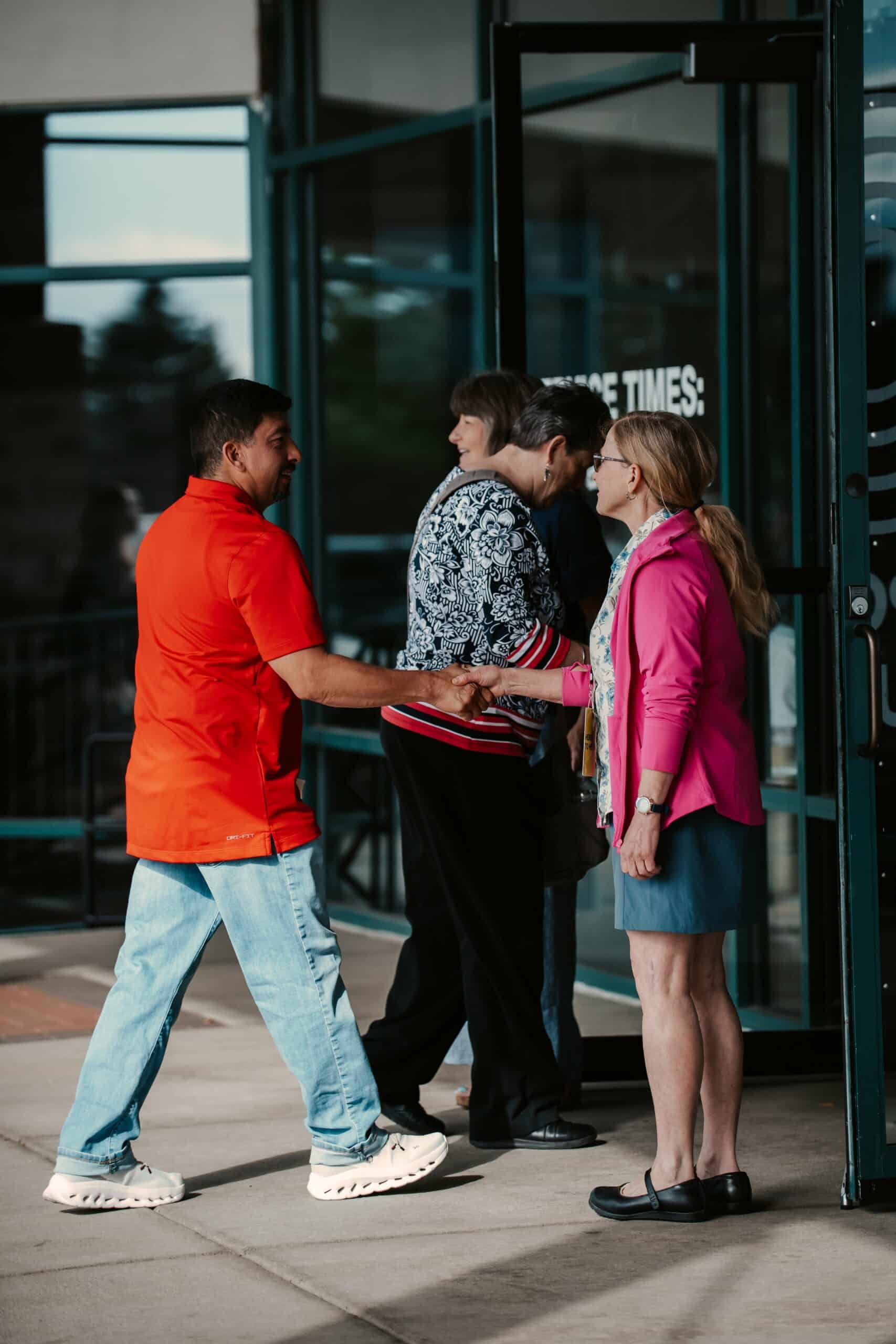 A man in a bright orange shirt shakes hands with a woman at an entrance while another woman in a patterned sweater and an older woman walk into a building. The entrance features glass doors and a welcome sign.