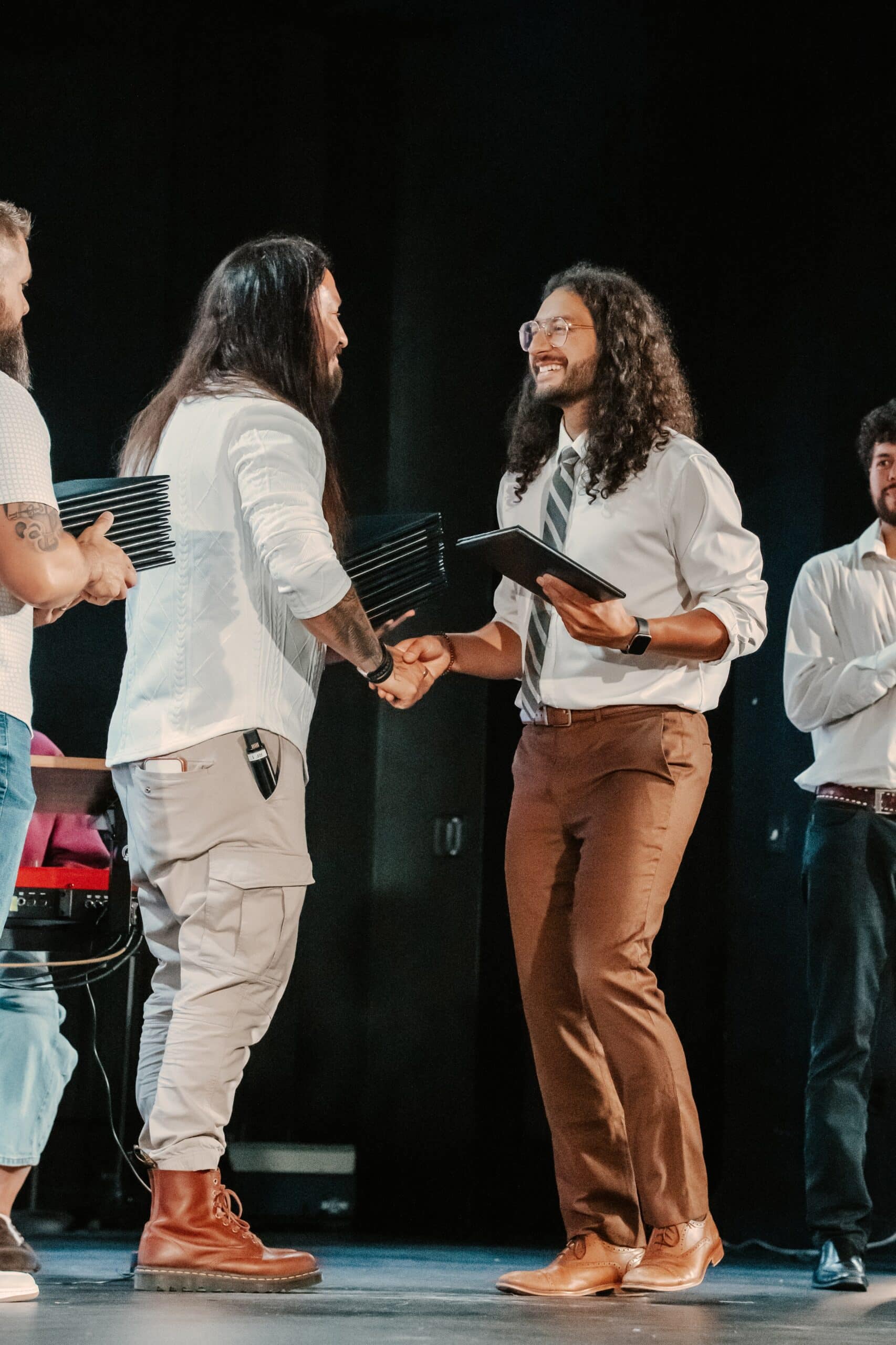 Two men are shaking hands on stage, with one wearing a white shirt and brown tie while receiving a certificate from the other in a white sweater and cargo pants. In the background, additional individuals in formal attire are visible.