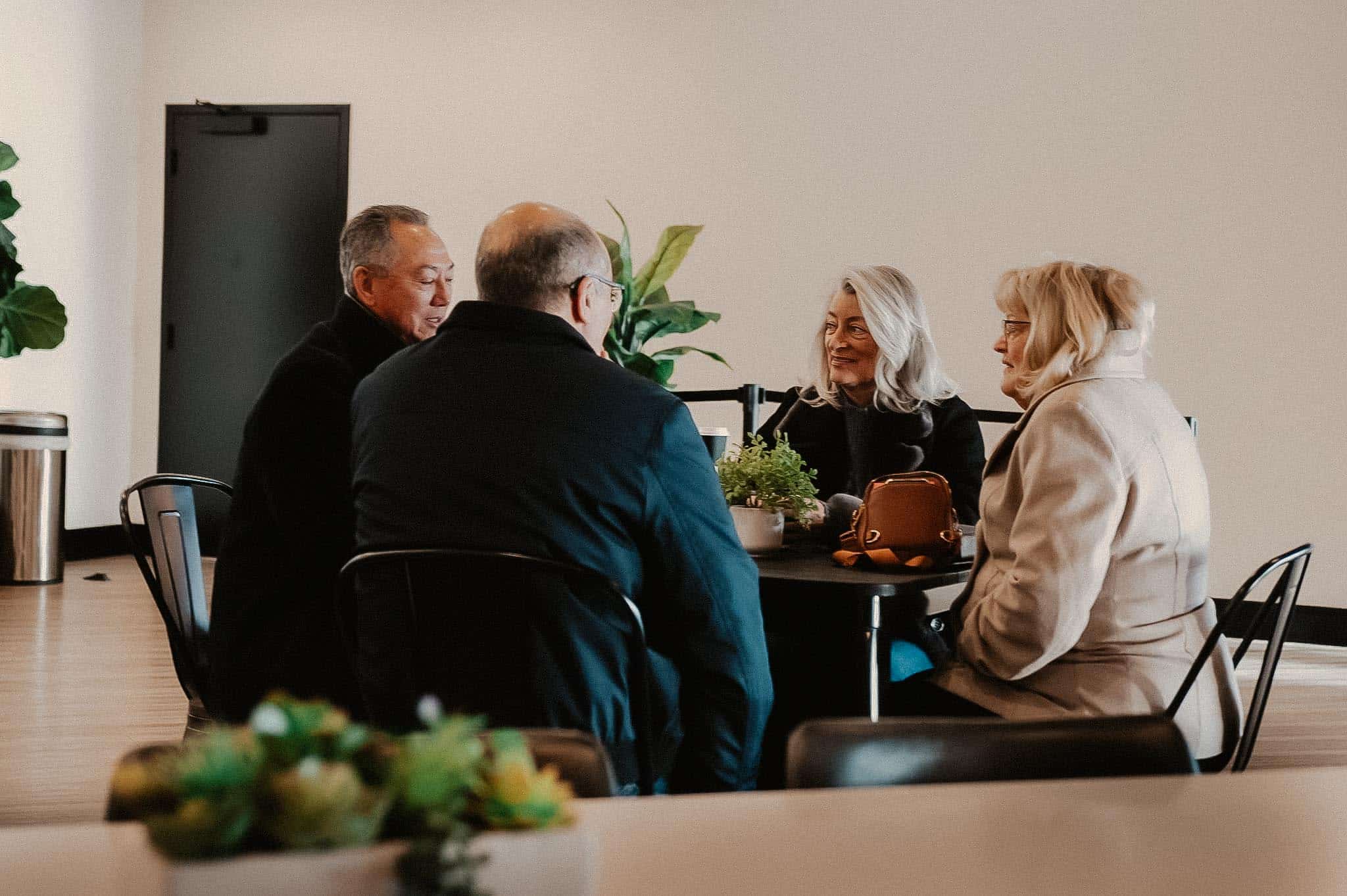 A group of four adults are sitting around a small black table in a modern indoor space. Two women and two men are engaged in conversation, with potted plants visible nearby and a trash bin in the background.