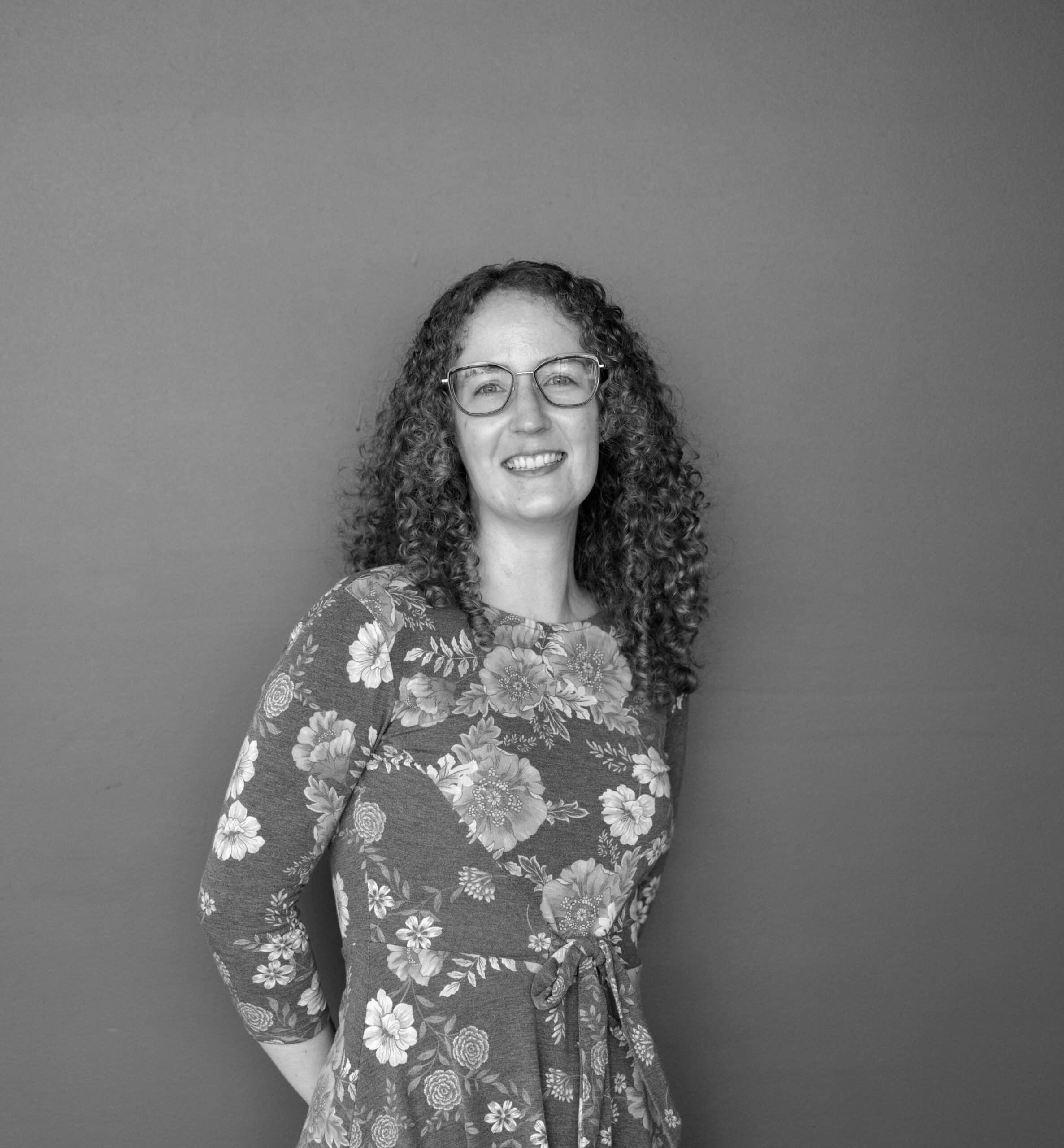 A woman with curly hair wearing glasses stands against a plain gray background. She is dressed in a floral-patterned dress with three-quarter sleeves and has her hand resting on her hip.