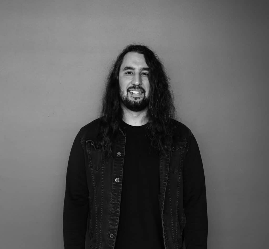 A man with long hair stands against a plain gray background. He is wearing a black T-shirt and a denim vest, with a slight smile visible.