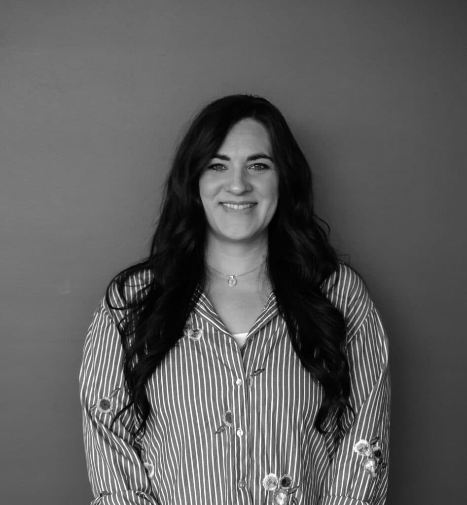 A woman with long dark hair is standing against a gray background. She is wearing a striped shirt with floral embroidery and a necklace. The photo is styled in black and white, emphasizing her outfit and features.