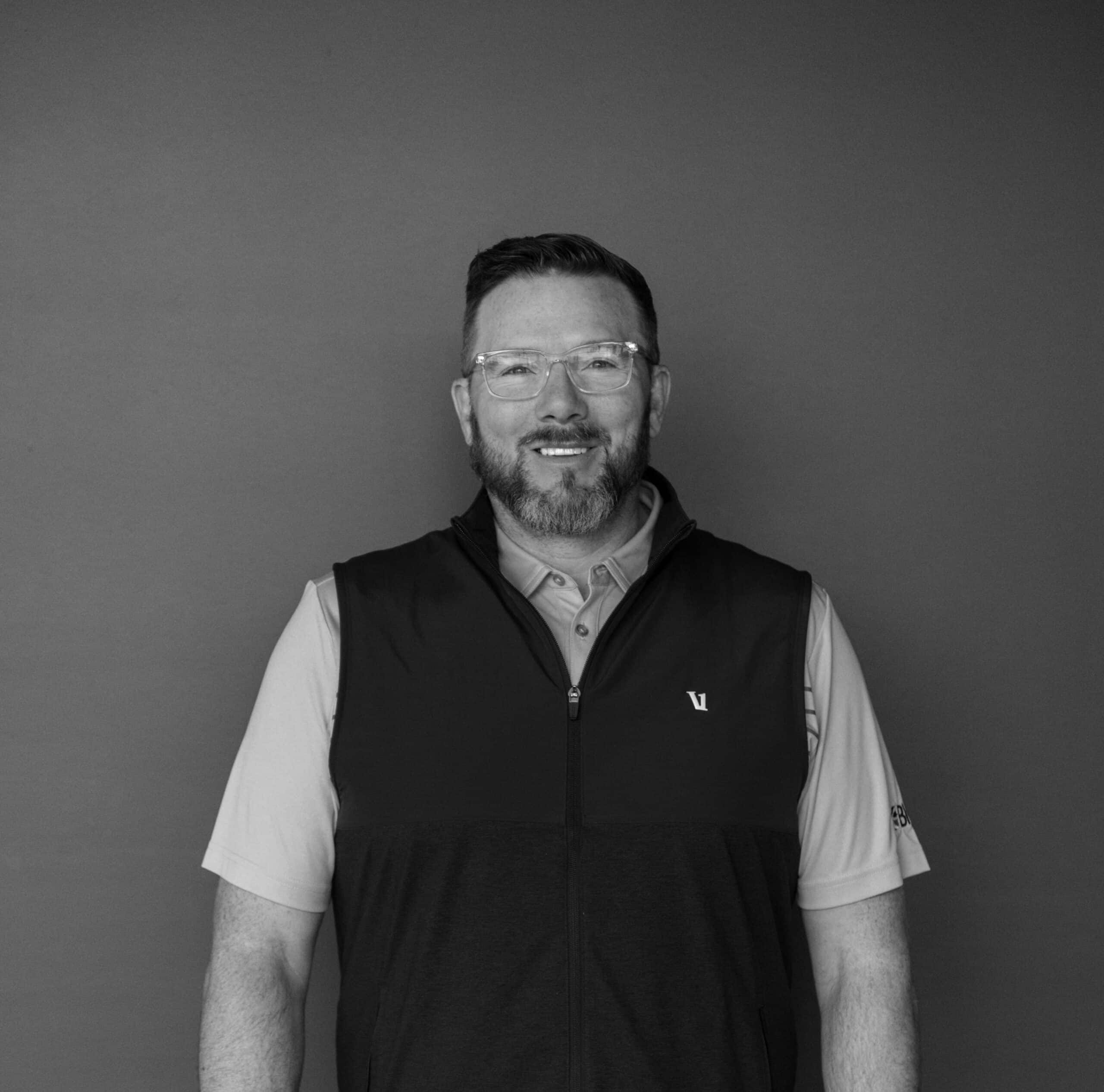 A man with glasses and a beard wears a dark vest over a light collared shirt. He is standing in front of a solid gray background, smiling towards the camera. The image is in black and white.