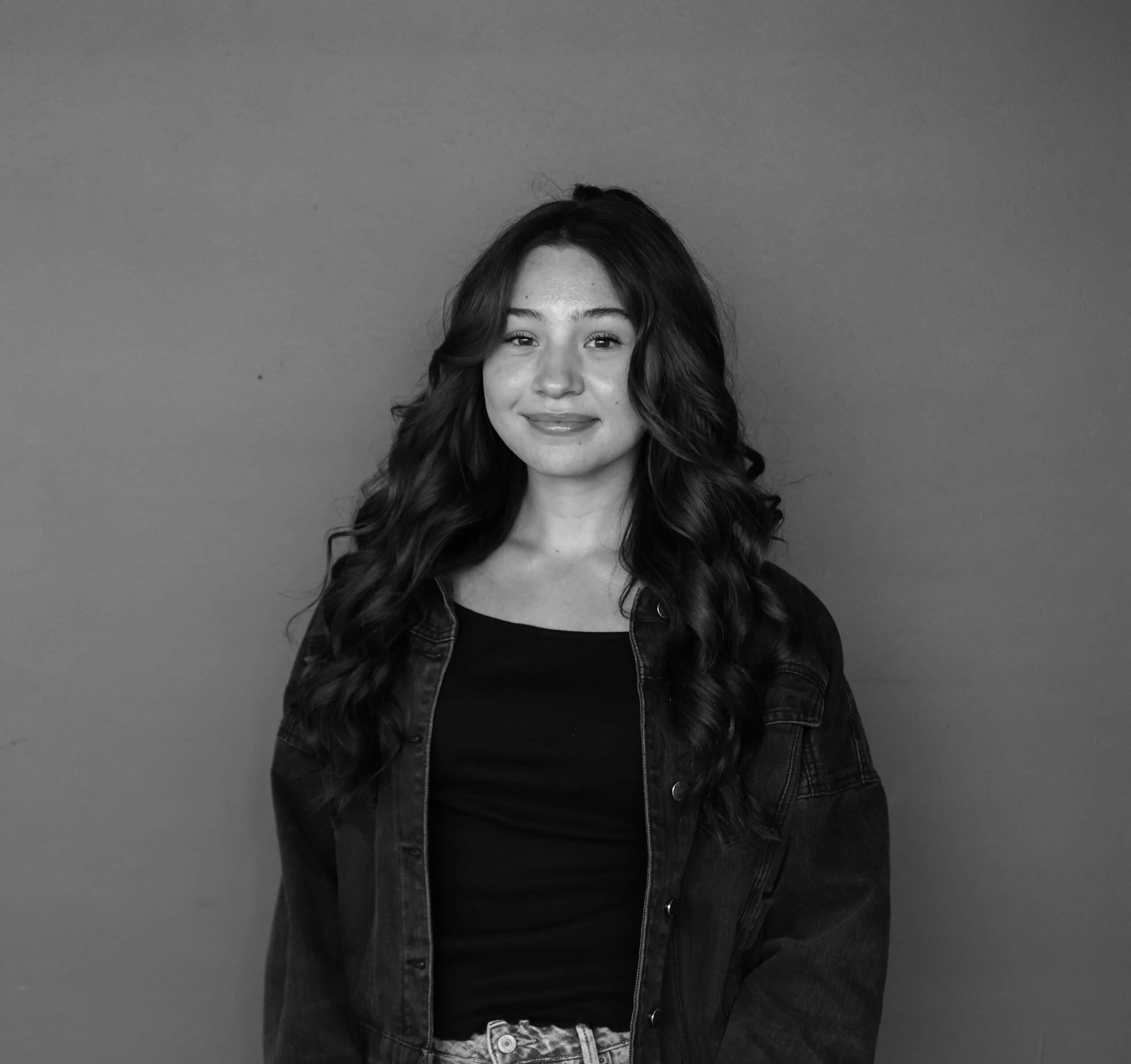 A young woman with long, wavy hair stands against a plain background, wearing a black top and an oversized denim jacket. The image is in black and white, showcasing her relaxed pose and the details of her outfit.