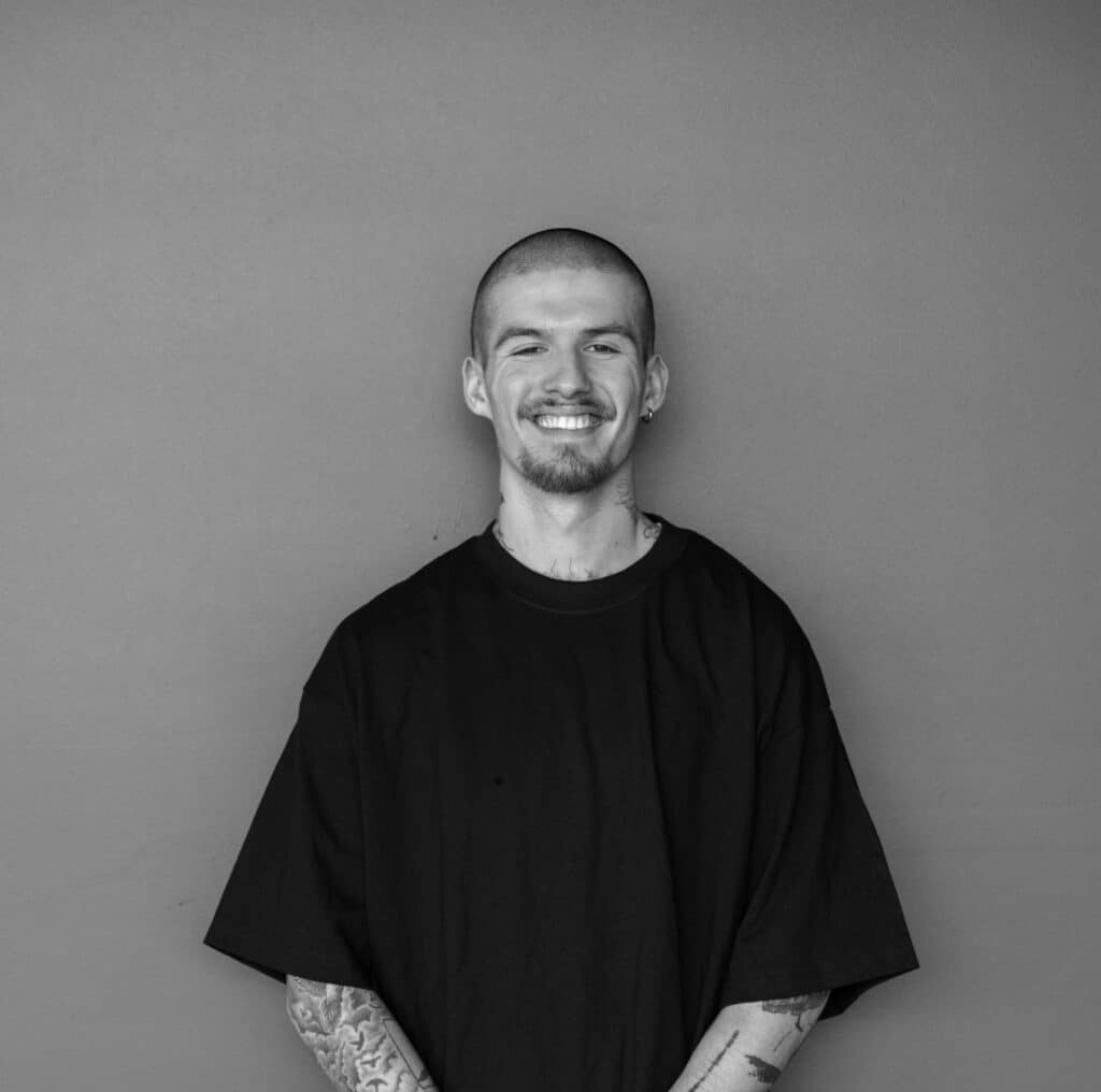 A man with a shaved head stands smiling in front of a plain gray wall. He is wearing a loose-fitting black t-shirt, and his arms are partially visible, showing tattoos.
