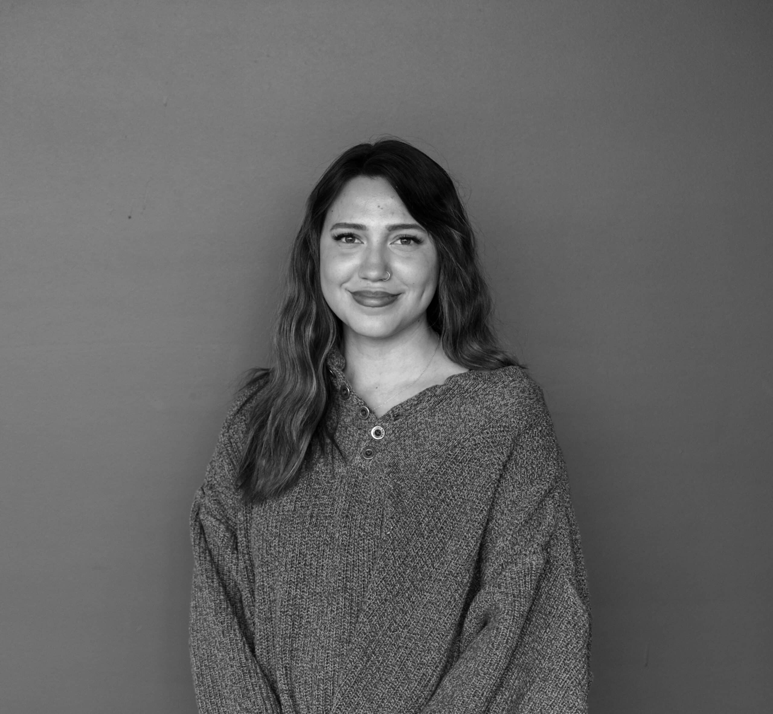 A young woman with long, wavy hair is smiling while wearing a gray, oversized sweater. She stands against a plain gray background, with her hands relaxed in front of her.