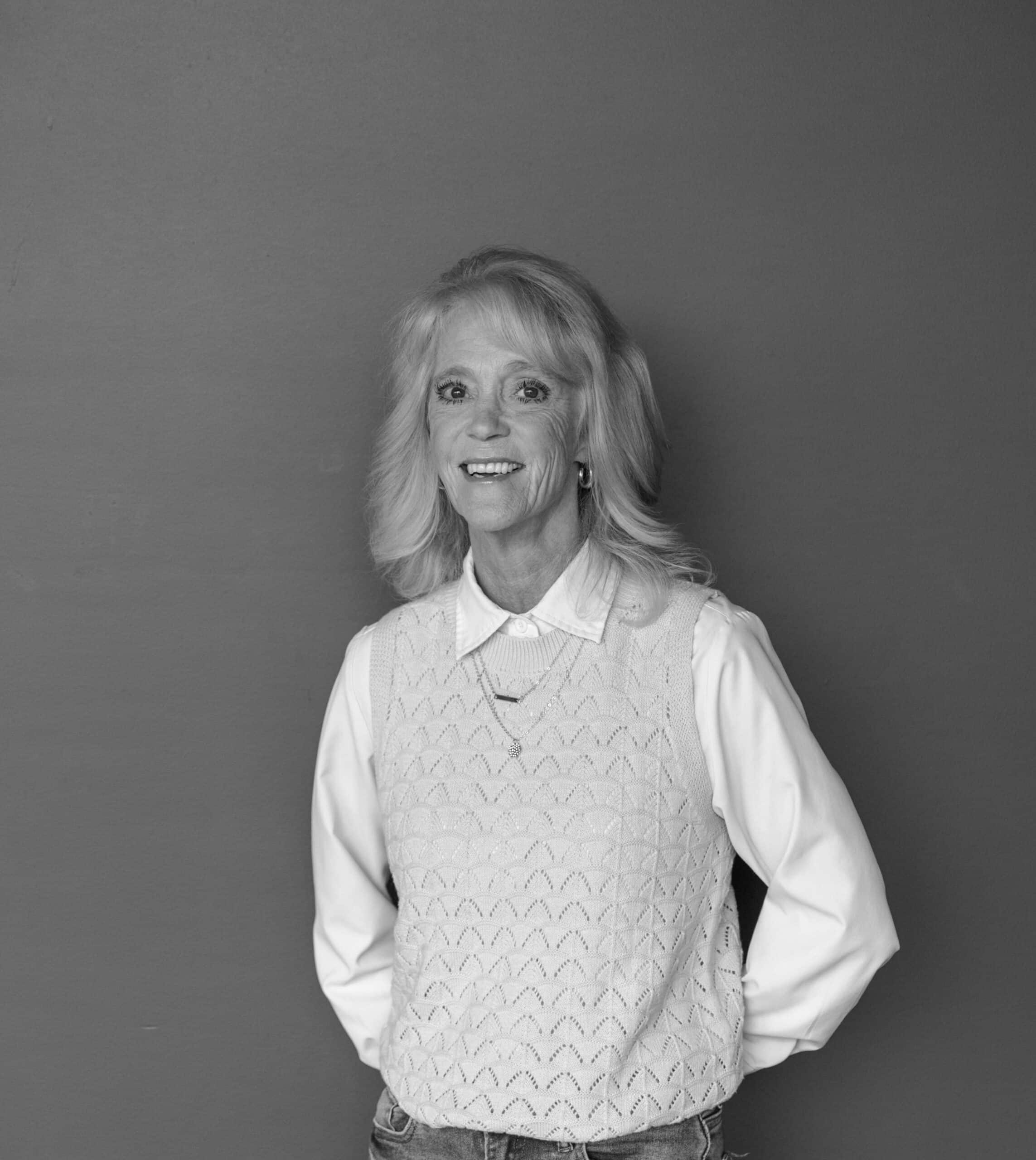 A woman with long, light-colored hair stands against a gray wall. She wears a white collared shirt under a light knit sweater, with her hands resting on her hips.