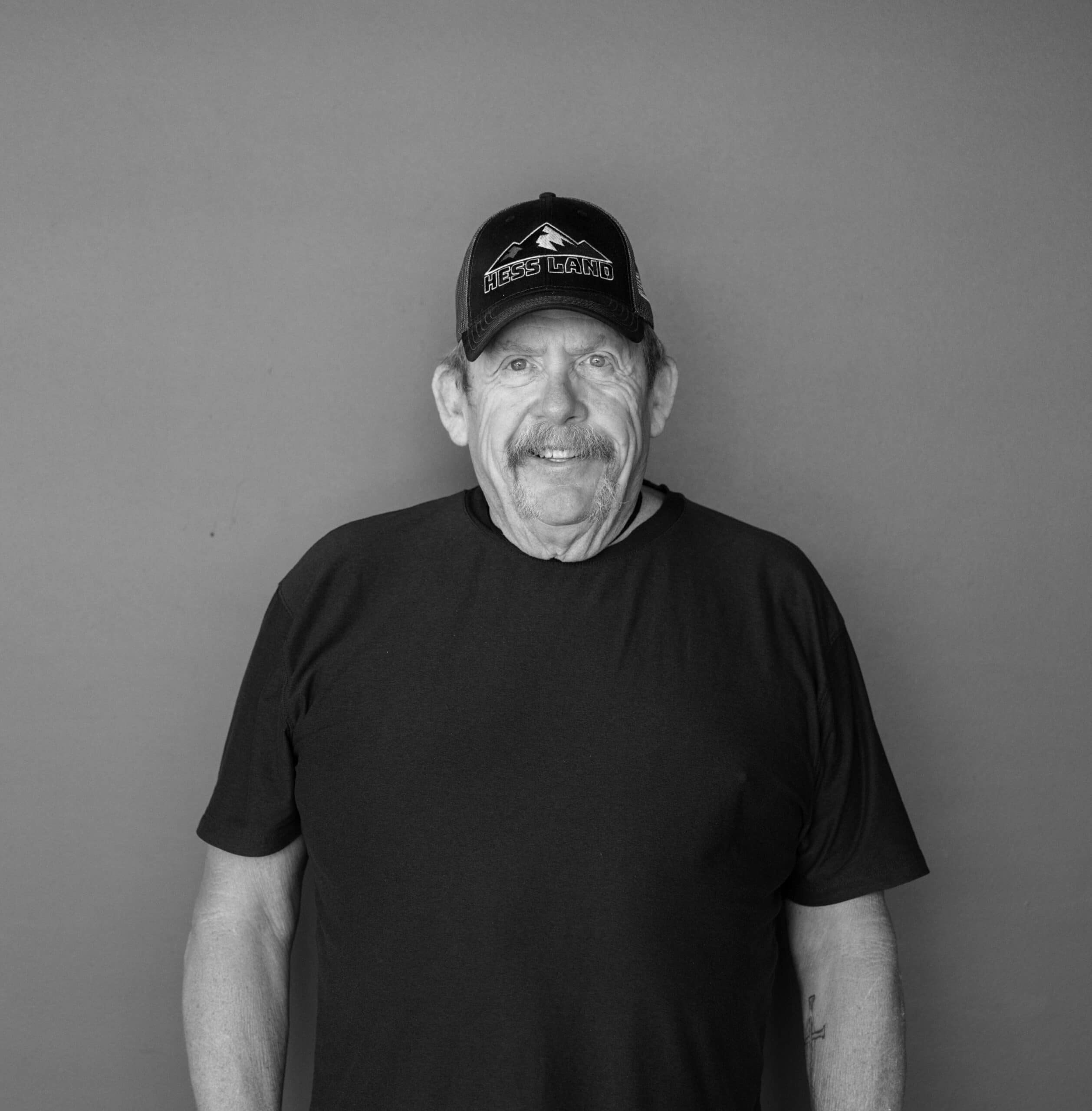 A man with a mustache wearing a black t-shirt and a black cap that reads "Less Land" stands against a plain gray background. He appears to be in a relaxed pose with his arms at his sides.