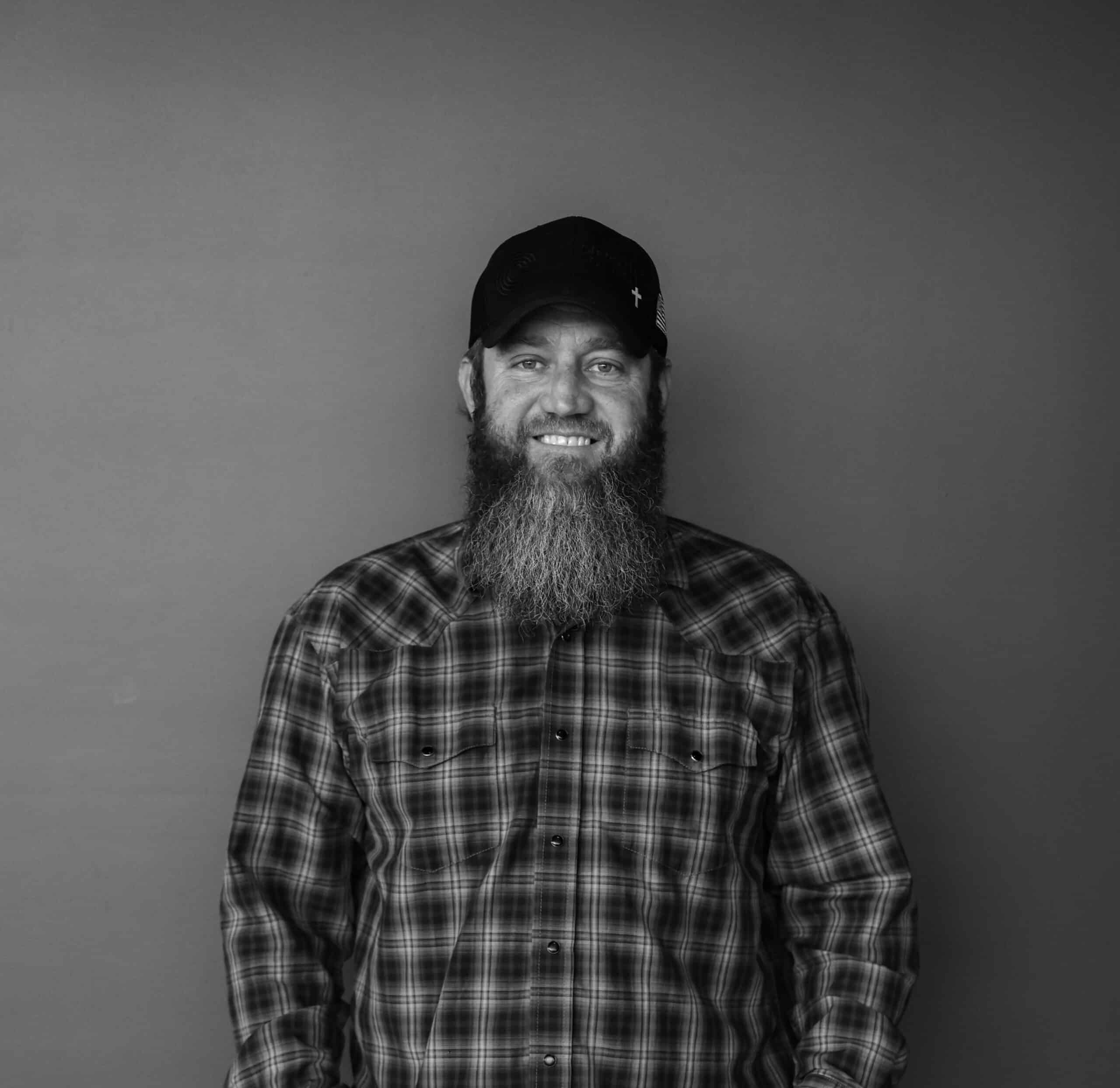 A man with a long beard wearing a black cap and a plaid shirt stands against a plain background. His posture is straight, and he faces the camera directly.