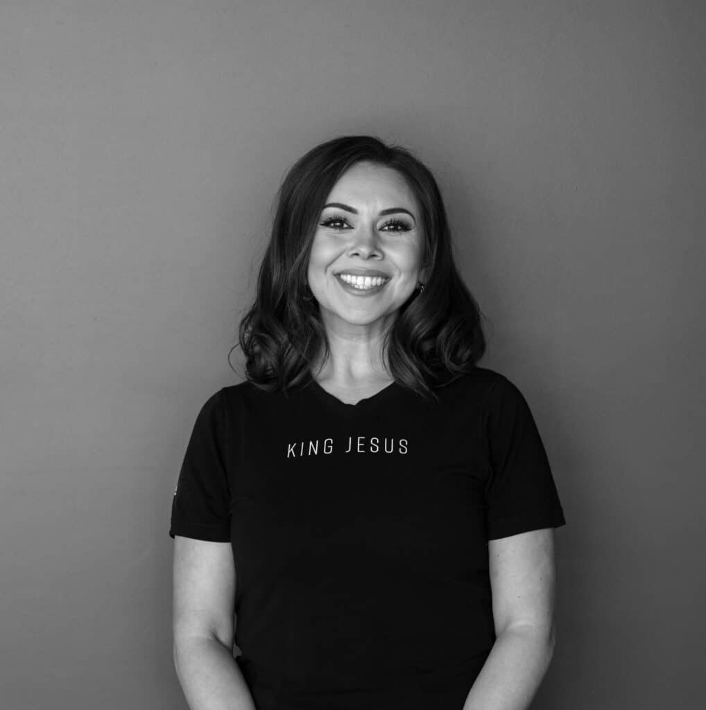 A woman with shoulder-length brown hair wearing a black t-shirt with the text "KING JESUS" stands against a gray background. She is positioned centrally and smiles at the camera with arms relaxed at her sides.