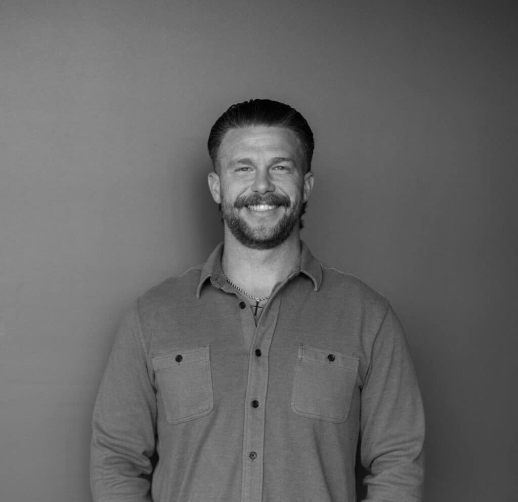 A man with short hair and a mustache stands in front of a solid gray backdrop. He is wearing a button-up shirt with the sleeves rolled up and has a small cross necklace. The image is in black and white.
